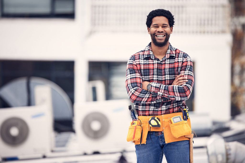 A smiling, professional HVAC technician wearing a tool belt standing in front of outdoor air conditioning units, featured on Air Alliance Heating & Cooling's guide on how to choose the right HVAC contractor in Cottleville, MO, for summer AC repairs.